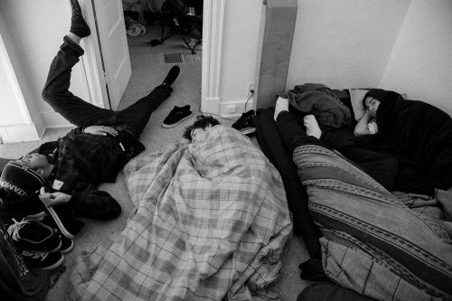 Black and white photo of four people sleeping indoors on bedding and the floor, along with blankets, shoes, and various objects.