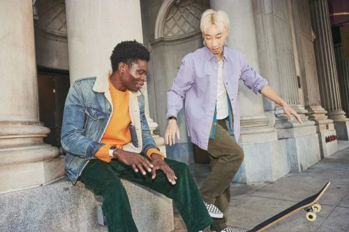 Two men, one sitting on concrete, the other standing near a skateboard, dressed in casual wear against a backdrop of stone columns.