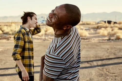 Black man laughing outdoors in a striped t-shirt with another man standing in the background.