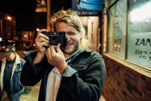 Man with blond hair takes a picture with a Leica camera in an urban setting at night, illuminated by streetlights.