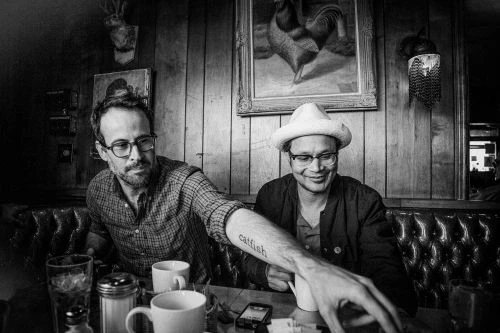 Two men with glasses sit in a restaurant, one has a tattoo on his arm. Cups and a sugar dispenser are visible on the table.