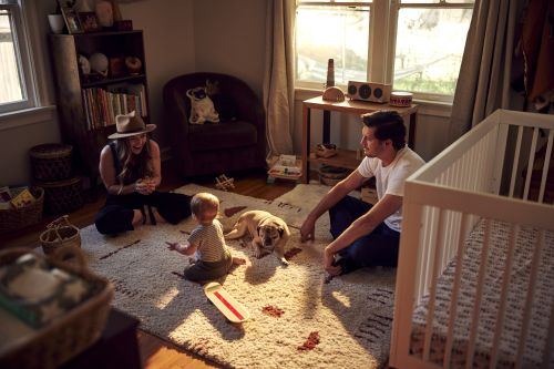 Indoor lifestyle scene with a family of three—mother, father, and child—sitting on a rug with their pug dog and a skateboard, bathed in natural light.