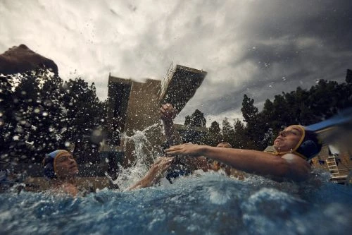 Water polo action shot: players compete in a pool with water splashing, with a cloudy sky and buildings in the background.