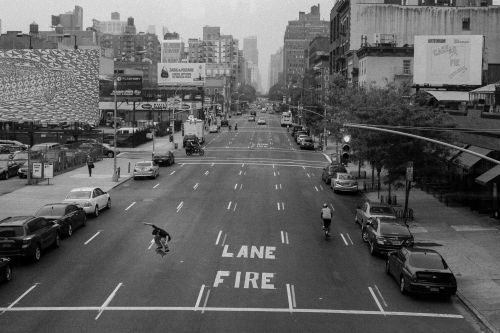 Black and white image of a skateboarder mid-air on a city street, with cars and buildings in the background, and the words 'LANE FIRE' visible on the road.