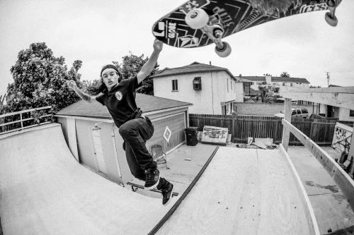 A black and white outdoor action shot of a skateboarder performing a trick in a backyard skate park, captured with a fisheye lens.