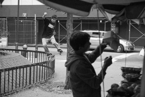 Black and white shot of a young man skateboarding on a railing, with another man in the foreground, and a city street scene background.