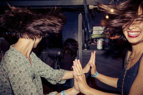 Two adult women with windblown hair clap hands, smiling joyfully in an indoor setting, showcasing fashion and active lifestyle elements.