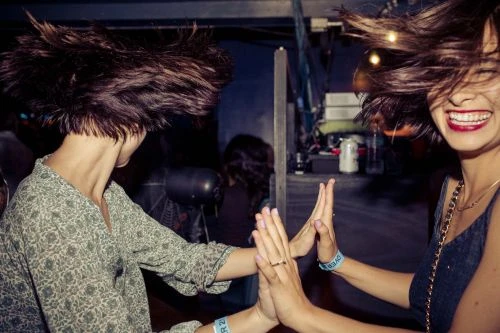 Two adult women with windblown hair clap hands, smiling joyfully in an indoor setting, showcasing fashion and active lifestyle elements.