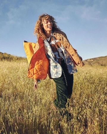 Smiling woman with curly hair in a jacket and patterned shirt stands in a sunlit field of tall golden grass in a rural setting.