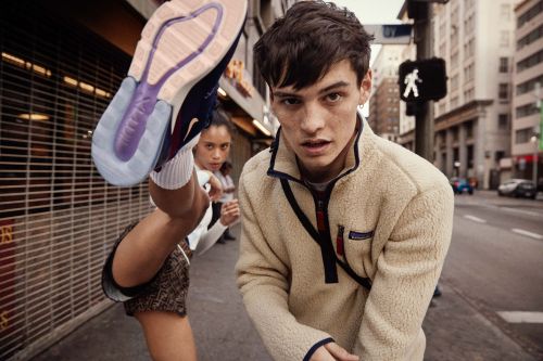 Two athletic people pose in an urban setting, one lifting a leg to display a purple-soled Nike Air Max 270 sneaker, beige fleece jacket.
