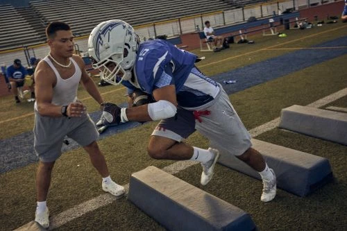 Two male athletes practicing football drills on a grass field under natural lighting, wearing athletic gear and helmets.