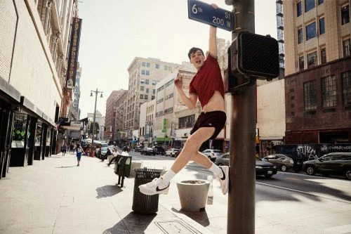 Young man hangs from traffic light pole in urban street; athletic pose, sportswear, white sneakers, 6th Street signage, sunny day, buildings.