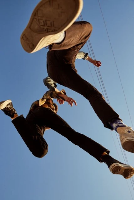 Two young adults in mid-air wearing brown pants and Converse shoes, photographed from a low angle against a clear blue sky.