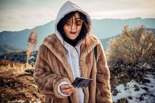 Woman in fur coat with sunglasses stands in a sunny, snowy landscape with mountains, looking at an object in her hand with surprise.