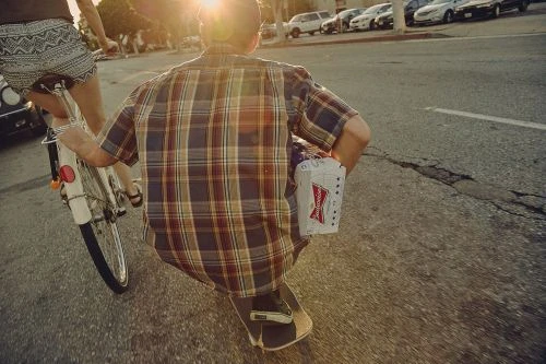 A man riding a skateboard next to a person on a bicycle on an asphalt street with parked cars in the background.