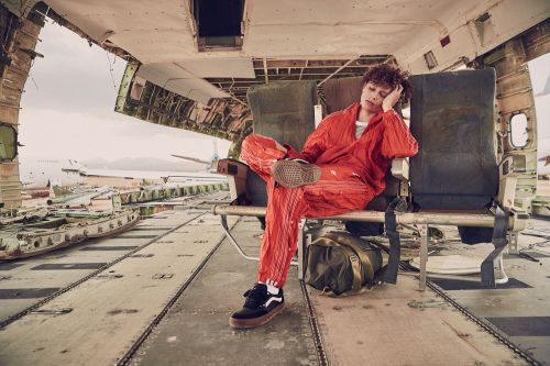 Person in an orange jumpsuit sits in an airplane seat inside of an abandoned plane. Travel bag is on the floor and shoes visible.
