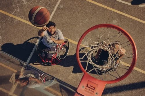 A male athlete in a wheelchair plays basketball, showcasing athletic skill and determination on an outdoor court, viewed from a high angle.