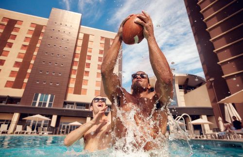 Two athletic men are playing with a football in an outdoor swimming pool in front of a large building on a sunny day with blue skies.