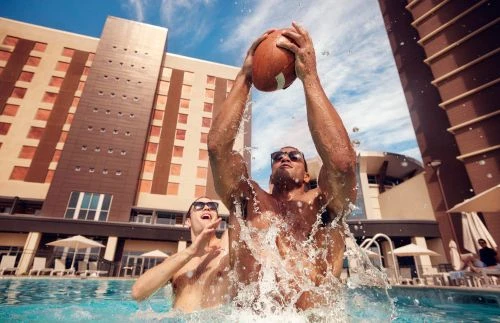 Two athletic men are playing with a football in an outdoor swimming pool in front of a large building on a sunny day with blue skies.