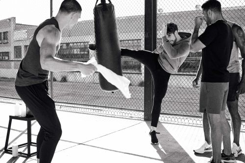 Group of four adult men in athletic wear training outdoors with a punching bag in black and white; one man is kicking the bag.