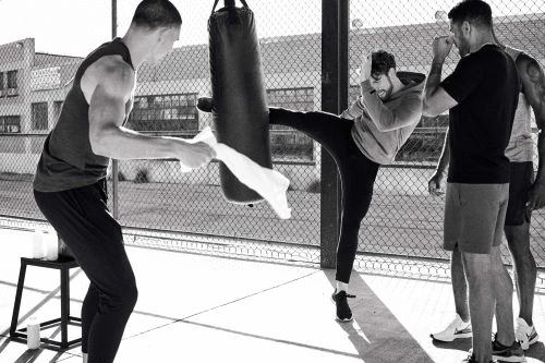 Group of four adult men in athletic wear training outdoors with a punching bag in black and white; one man is kicking the bag.