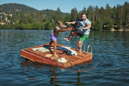 Two men on a wooden raft in a lake with forest hills in the background, one helping the other regain balance. Summer fun concept.