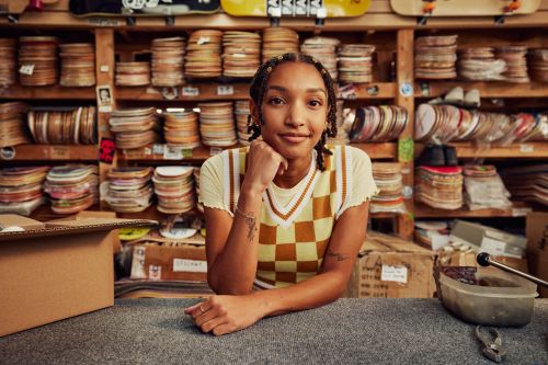 Young adult with braids poses behind a retail counter with shelves of skateboards and accessories in a warm-toned indoor setting.