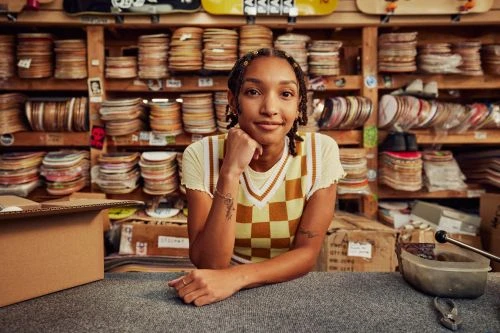 Young adult with braids poses behind a retail counter with shelves of skateboards and accessories in a warm-toned indoor setting.