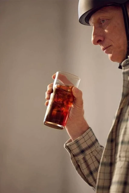 Older man in a helmet holds a glass of iced tea, looking down, wearing a plaid shirt in a neutral indoor setting with soft lighting.