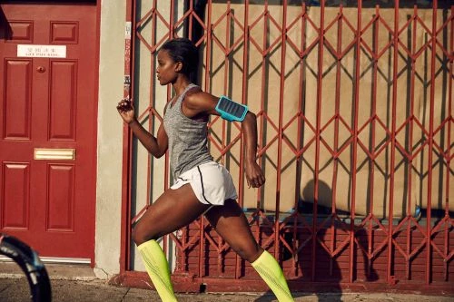 Athletic woman running outdoors wearing workout gear, phone armband, and compression socks, with a red door and gate in the background.