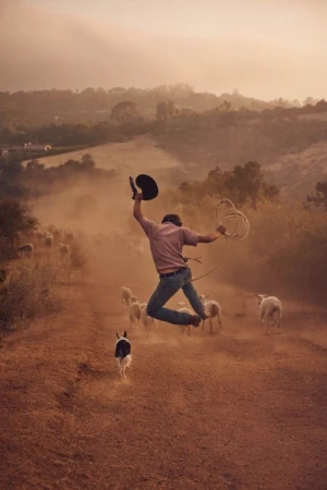Man jumps mid-air holding cowboy hat and lasso, with a dog and herd of sheep on a dusty path, set against a sunlit, rural landscape.