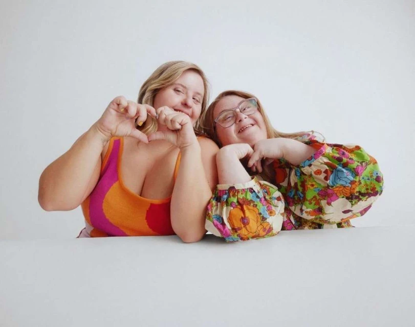 Two women smiling and making a heart shape with their hands. One woman is wearing glasses and a floral shirt. Indoor setting.