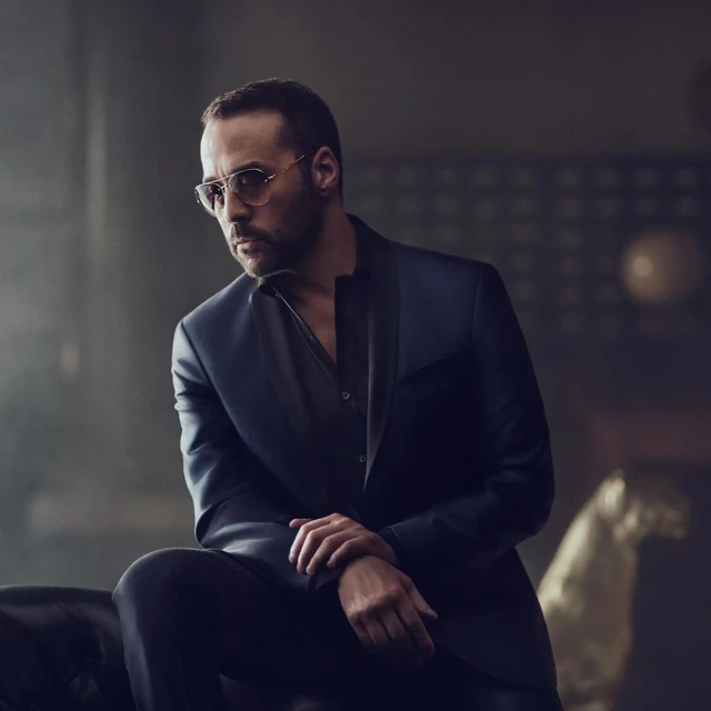 Portrait of a man in a navy suit and glasses, seated on a couch in a dimly lit room with a gold-colored decorative object.