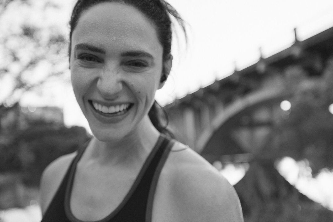 Close up of a smiling woman wearing a tank top with a blurred bridge and trees in the background. Black and white photograph.