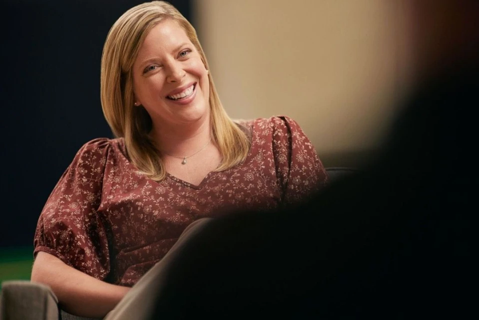Smiling adult woman with blonde hair and wearing a floral print blouse sitting and posing in front of a light colored wall.