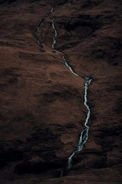 A long exposure captures a stream flowing down a hillside, showcasing blurred water motion and a dark, earthy-toned landscape.