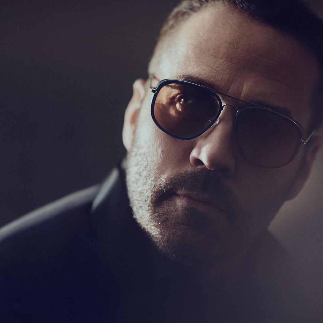 Close-up portrait of a man wearing tinted aviator sunglasses and a dark blazer in a softly lit, high contrast studio setting.