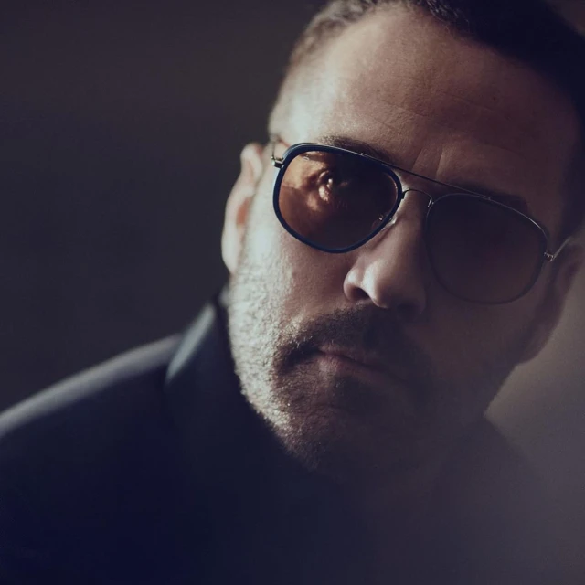 Close-up portrait of a man wearing tinted aviator sunglasses and a dark blazer in a softly lit, high contrast studio setting.