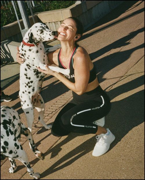 Woman in athletic wear smiles and pets a Dalmatian dog outdoors on a sunny day near a railing, showcasing active lifestyle.