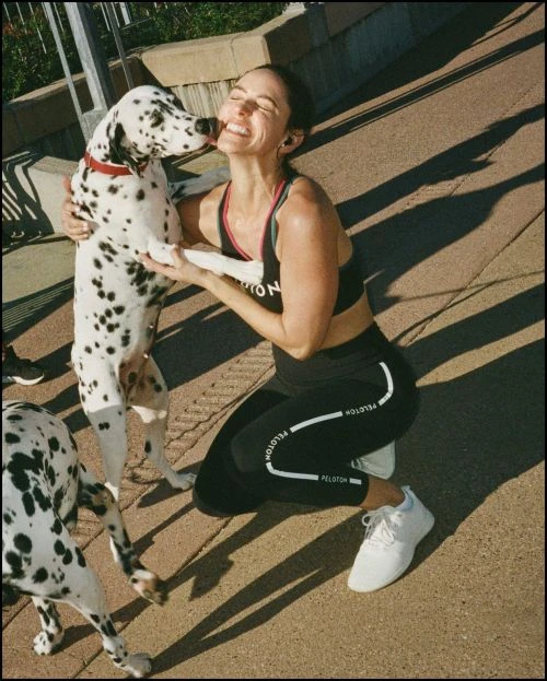 Woman in athletic wear smiles and pets a Dalmatian dog outdoors on a sunny day near a railing, showcasing active lifestyle.