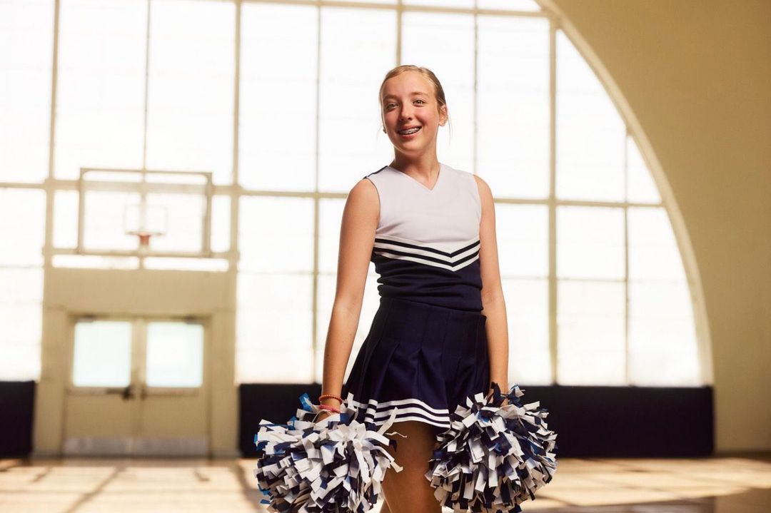 Teen cheerleader in uniform smiles, holding pom-poms in gymnasium. Navy and white uniform, braces, and natural lighting.
