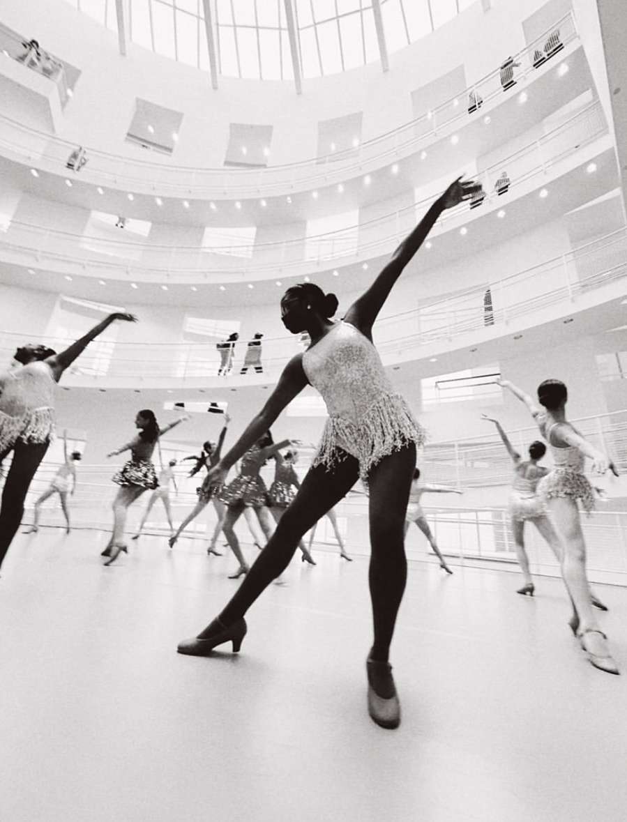 A group of dancers in white dresses performing in a large room with a skylight and multi-level balconies in a black and white photograph.