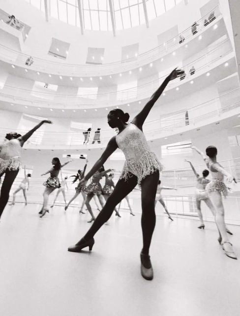 A group of dancers in white dresses performing in a large room with a skylight and multi-level balconies in a black and white photograph.
