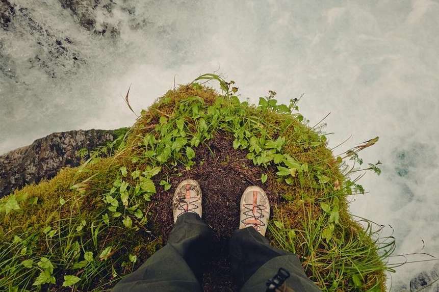 Person standing on mossy cliff edge with view of rushing whitewater river, wearing outdoor trousers and hiking shoes; scenic nature.