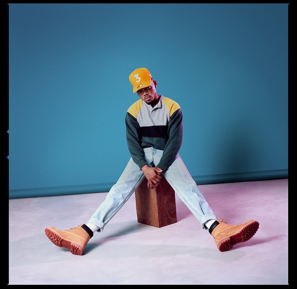 Young adult male in a yellow baseball cap and Timberland boots sits with his legs apart against a blue backdrop.