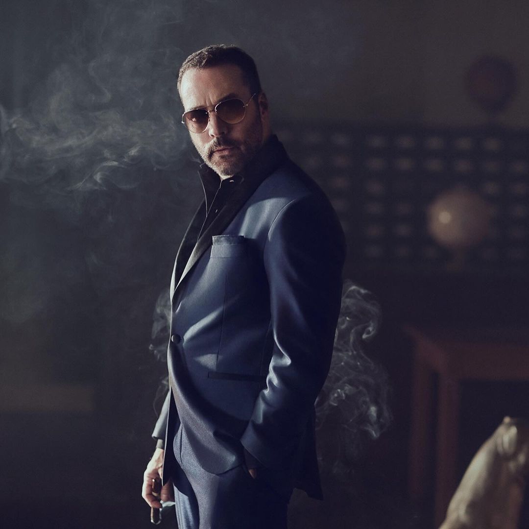 Man wearing a blue blazer and sunglasses standing amidst swirling smoke in a dimly lit interior setting.