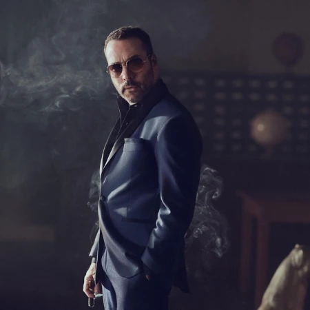 Man wearing a blue blazer and sunglasses standing amidst swirling smoke in a dimly lit interior setting.