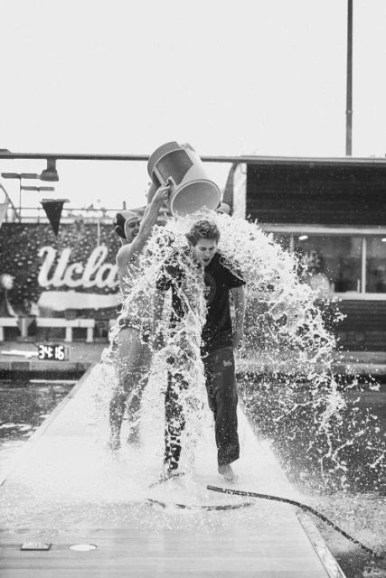 Two people dousing a third person with water from a bucket near a pool; a monochrome image capturing action and splash effects.