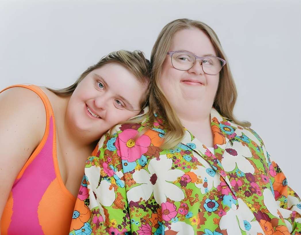 Two women with Down syndrome pose closely in a studio shot. One wears glasses and a floral blouse, the other a bright swimsuit.