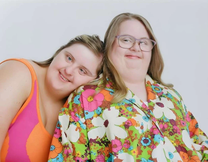 Two women with Down syndrome pose closely in a studio shot. One wears glasses and a floral blouse, the other a bright swimsuit.